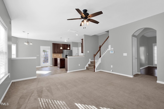 unfurnished living room with arched walkways, light carpet, ceiling fan with notable chandelier, baseboards, and stairway