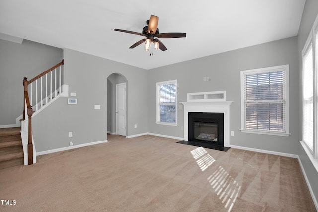 unfurnished living room with arched walkways, light colored carpet, a fireplace with flush hearth, baseboards, and stairs