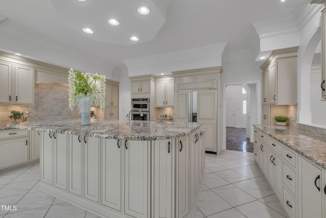 kitchen featuring arched walkways, cream cabinets, light stone countertops, an island with sink, and crown molding