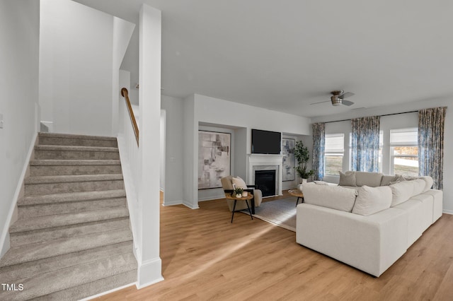 living area with ceiling fan, light wood-style flooring, baseboards, stairway, and a glass covered fireplace