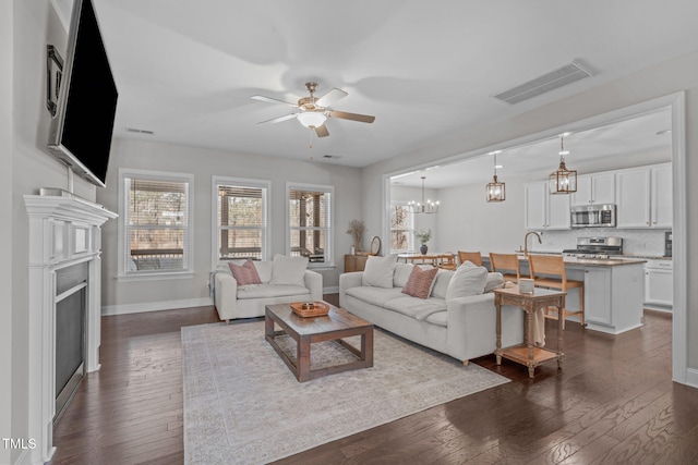 living area featuring ceiling fan with notable chandelier, dark wood finished floors, visible vents, and baseboards