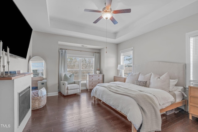 bedroom featuring a ceiling fan, dark wood finished floors, baseboards, a raised ceiling, and a glass covered fireplace