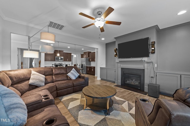 living area with visible vents, light wood-style flooring, ornamental molding, a glass covered fireplace, and a ceiling fan