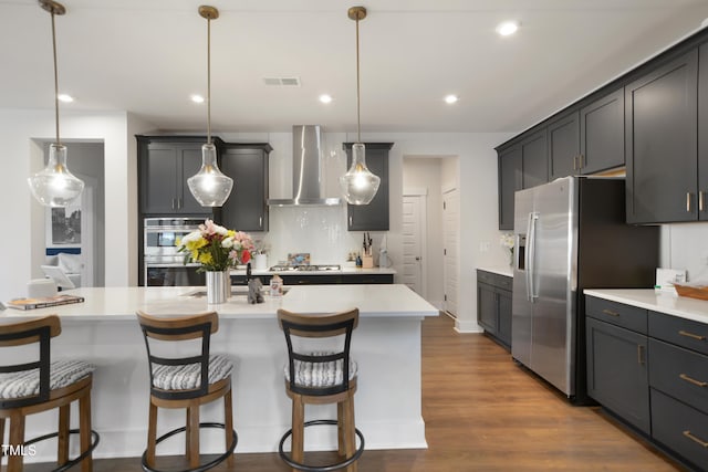 kitchen with dark wood-style floors, wall chimney exhaust hood, light countertops, appliances with stainless steel finishes, and a kitchen bar