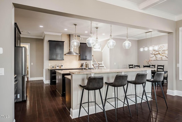 kitchen with a breakfast bar area, a center island with sink, freestanding refrigerator, crown molding, and wall chimney exhaust hood