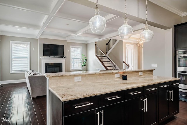 kitchen with dark wood finished floors, a glass covered fireplace, dark cabinetry, stainless steel double oven, and coffered ceiling