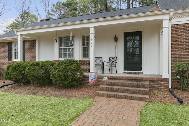 doorway to property featuring a shingled roof, covered porch, and brick siding