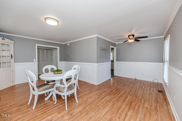 dining area with light wood finished floors, visible vents, a wainscoted wall, ornamental molding, and a ceiling fan