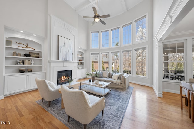 living area featuring light wood-type flooring, a ceiling fan, a glass covered fireplace, baseboards, and a towering ceiling