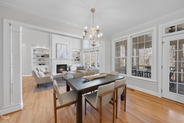 dining area with built in features, light wood-style floors, a glass covered fireplace, crown molding, and a notable chandelier