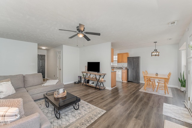 living area featuring a textured ceiling, ceiling fan with notable chandelier, visible vents, baseboards, and dark wood-style floors