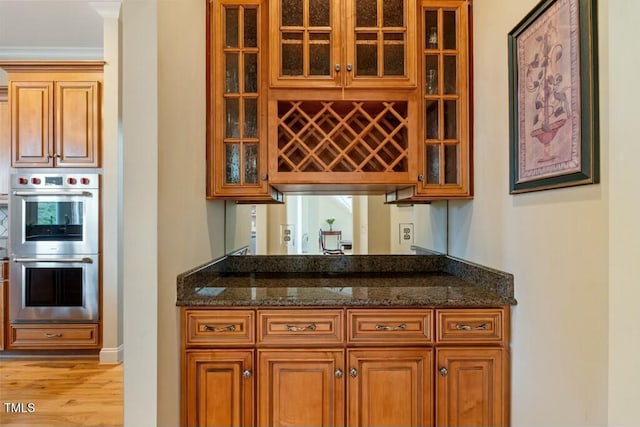 kitchen with brown cabinetry, stainless steel double oven, dark stone countertops, and glass insert cabinets