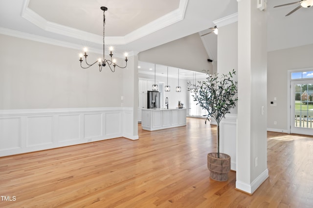 unfurnished dining area featuring a wainscoted wall, crown molding, a raised ceiling, light wood-style floors, and ceiling fan with notable chandelier