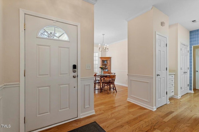 entryway with light wood finished floors, a wainscoted wall, an inviting chandelier, crown molding, and a decorative wall