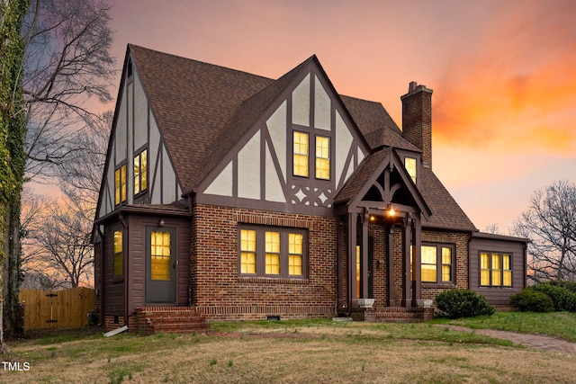 english style home featuring brick siding, a shingled roof, fence, stucco siding, and a chimney