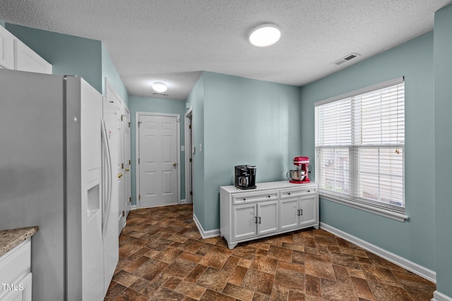 kitchen with white refrigerator with ice dispenser, white cabinetry, visible vents, baseboards, and stone finish flooring