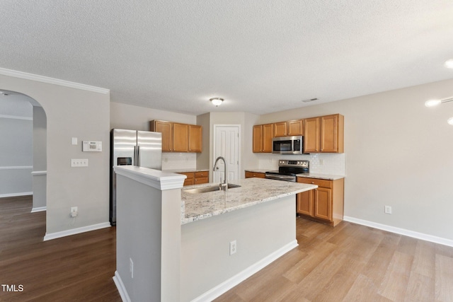 kitchen featuring arched walkways, light wood-style flooring, a kitchen island with sink, stainless steel appliances, and a sink