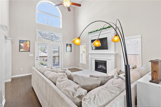 living room featuring ceiling fan, dark wood-style flooring, a fireplace, a towering ceiling, and baseboards