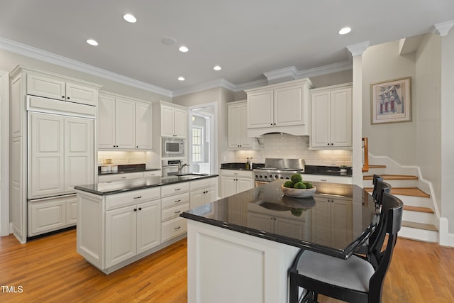 kitchen with light wood finished floors, dark countertops, built in appliances, a kitchen island with sink, and white cabinetry