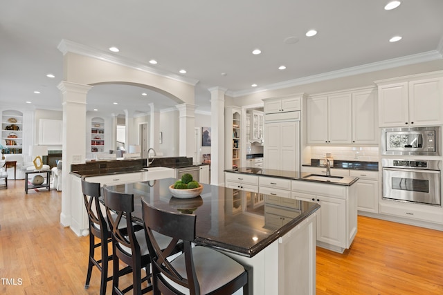 kitchen with light wood-style floors, an island with sink, stainless steel appliances, ornate columns, and a sink