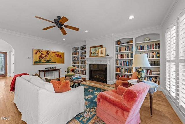 living room with wood finished floors, arched walkways, a fireplace, crown molding, and ceiling fan