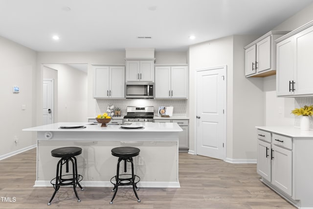 kitchen with light wood finished floors, an island with sink, appliances with stainless steel finishes, a breakfast bar, and backsplash