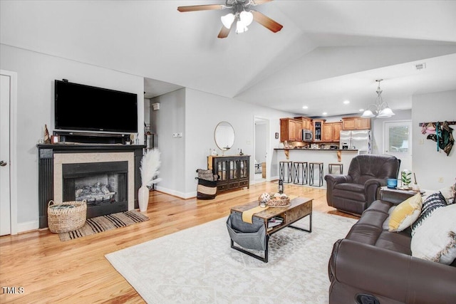 living area featuring baseboards, lofted ceiling, light wood-type flooring, a fireplace, and ceiling fan with notable chandelier