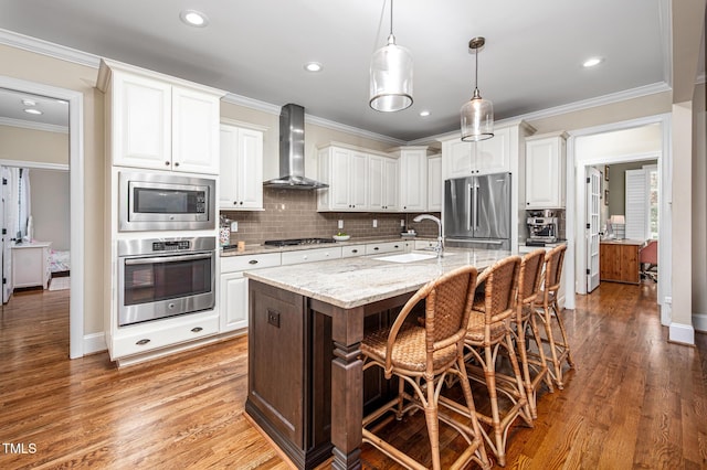 kitchen featuring appliances with stainless steel finishes, ornamental molding, white cabinetry, a sink, and wall chimney exhaust hood