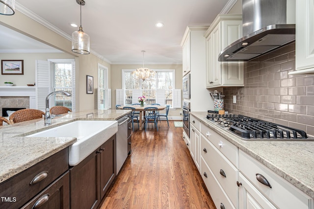 kitchen with appliances with stainless steel finishes, wood finished floors, dark brown cabinets, wall chimney range hood, and a sink