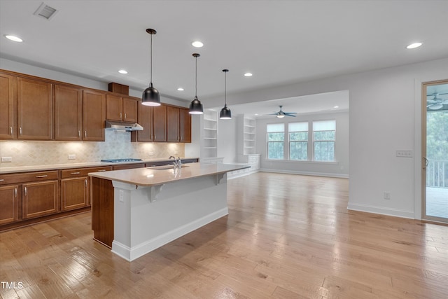 kitchen with under cabinet range hood, light wood-type flooring, brown cabinets, stainless steel gas stovetop, and a sink