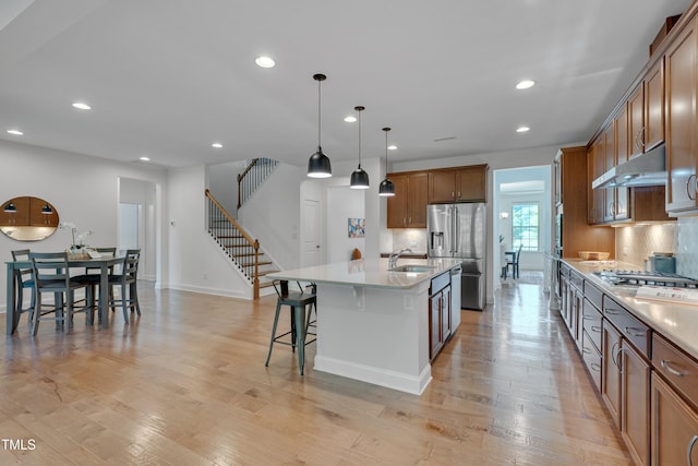 kitchen with a sink, brown cabinetry, a center island with sink, under cabinet range hood, and appliances with stainless steel finishes