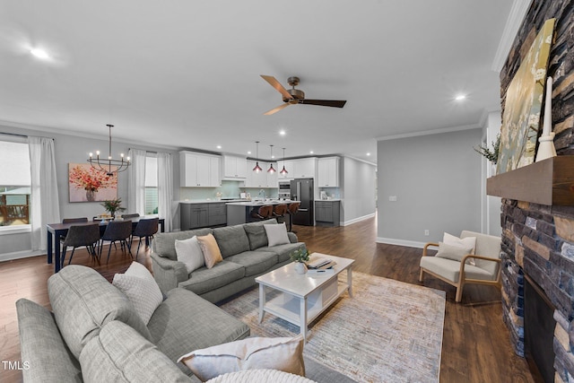 living area featuring dark wood-style flooring, ornamental molding, a stone fireplace, baseboards, and ceiling fan with notable chandelier