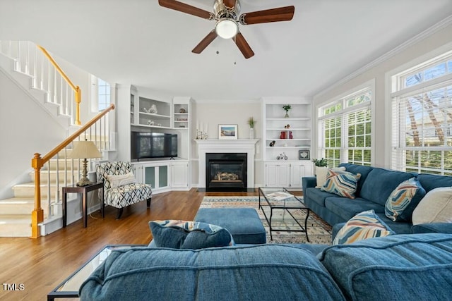 living area featuring ceiling fan, a fireplace with flush hearth, wood finished floors, stairs, and crown molding