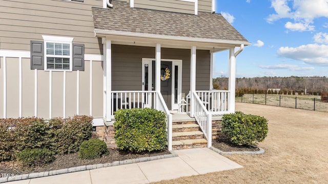 view of exterior entry featuring covered porch, a shingled roof, and fence