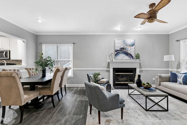 living room with ornamental molding, a glass covered fireplace, ceiling fan, wood finished floors, and baseboards