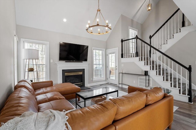 living area featuring a notable chandelier, a glass covered fireplace, wood finished floors, high vaulted ceiling, and stairs