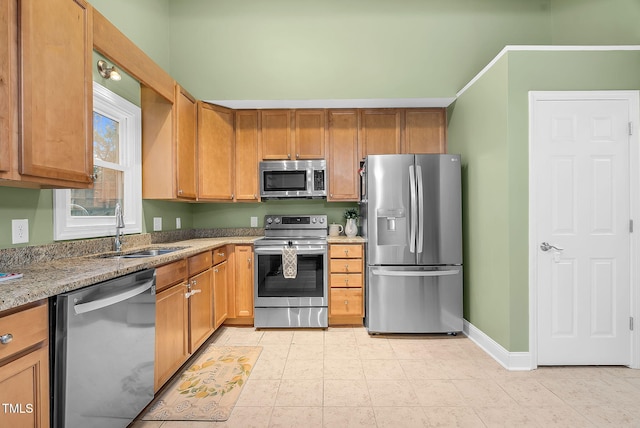 kitchen featuring light stone countertops, a sink, baseboards, appliances with stainless steel finishes, and brown cabinets