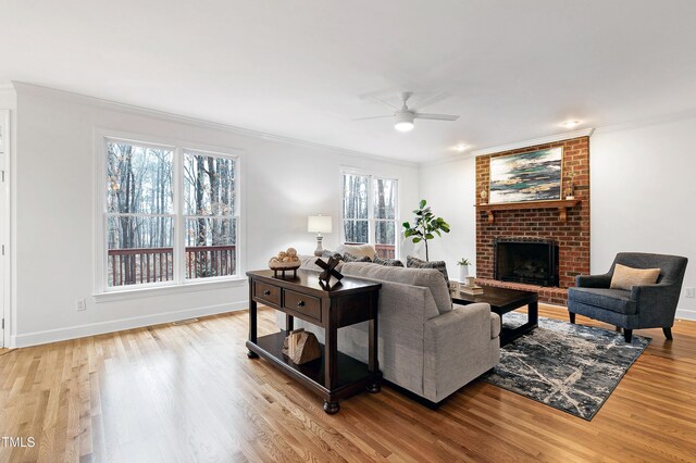 living room with ornamental molding, light wood-style floors, a brick fireplace, a ceiling fan, and baseboards