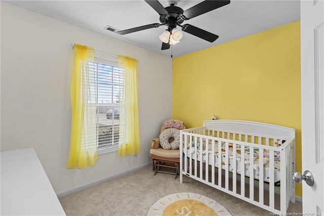 carpeted bedroom with a crib, baseboards, visible vents, and ceiling fan