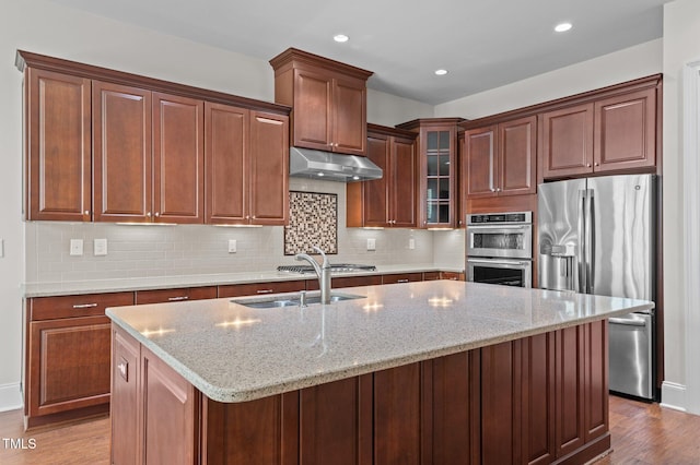 kitchen featuring wood finished floors, a center island with sink, a sink, stainless steel appliances, and under cabinet range hood