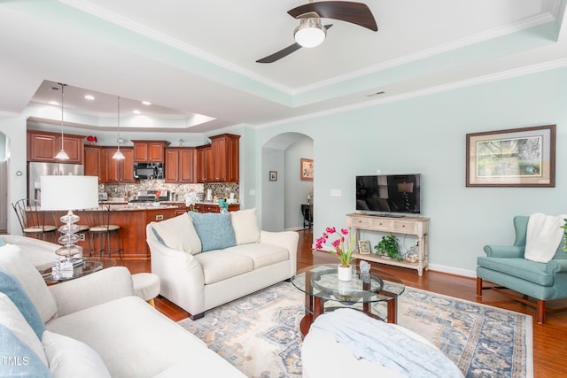 living room featuring arched walkways, ceiling fan, ornamental molding, wood finished floors, and a tray ceiling