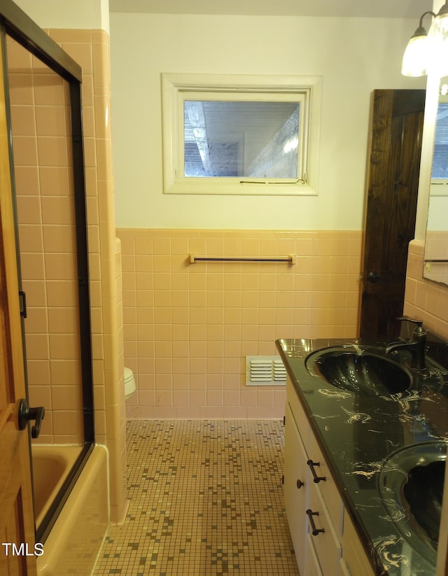 bathroom with tile walls, double vanity, bath / shower combo with glass door, visible vents, and a sink