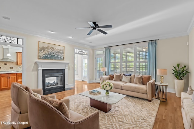 living room with light wood finished floors, baseboards, ornamental molding, a glass covered fireplace, and a ceiling fan