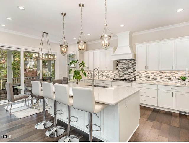 kitchen with a breakfast bar area, a kitchen island with sink, premium range hood, a sink, and tasteful backsplash