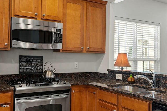 kitchen featuring brown cabinetry, appliances with stainless steel finishes, dark stone counters, and a sink