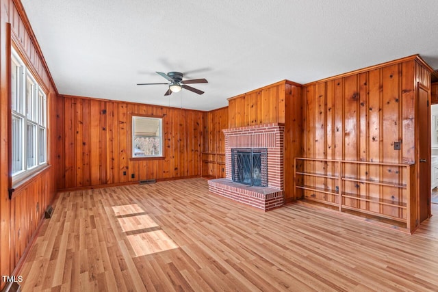 unfurnished living room featuring visible vents, a brick fireplace, ceiling fan, wooden walls, and light wood-type flooring