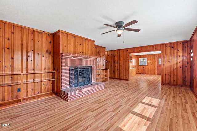 unfurnished living room featuring a brick fireplace, light wood-style flooring, a ceiling fan, and wood walls