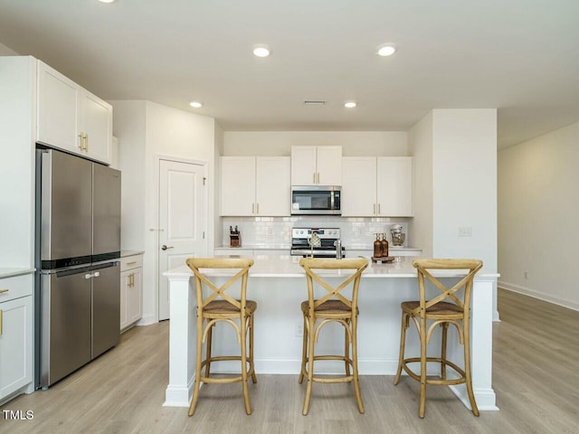 kitchen with a center island with sink, stainless steel appliances, light countertops, white cabinetry, and light wood-type flooring