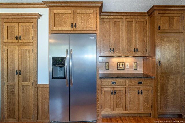 kitchen with backsplash, stainless steel fridge with ice dispenser, dark stone counters, wood finished floors, and brown cabinetry