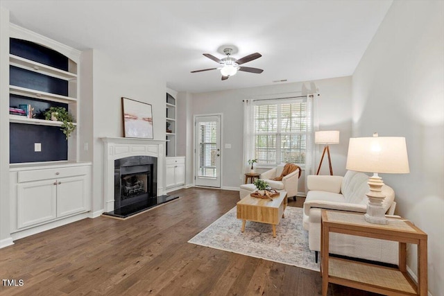 living room featuring baseboards, a ceiling fan, dark wood-type flooring, built in shelves, and a fireplace